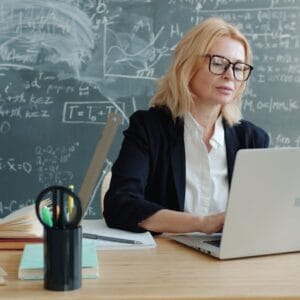 Teacher working on laptop in front of chalkboard.